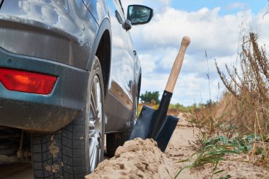 Yakınlarda bir kürek ile kumda sıkışmış, kullanıma hazır bir araba. Görüntü, arabanın tekerleğinin ve etrafındaki kumlu arazinin yakın çekimini yakalıyor. Bu da bir off-road macerasını ya da meydan okumayı gösteriyor..