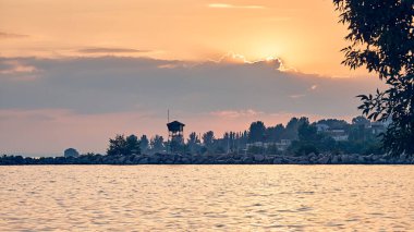 Güneş batan güneşin ılık renklerini yansıtan ağaçlar ve huzur dolu altın sularla çevrili, sahilde bir gözetleme kulesi silueti olan sakin bir su manzarasının üzerinde gün batımı..