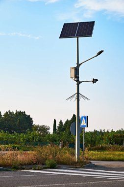 Sakin, kırsal bir bölgede yaya geçidinin yanında duran güneş enerjili bir sokak lambası. Sahne, modern teknolojinin ve doğal çevrenin bir karışımını parlak, berrak bir gökyüzü altında yakalıyor..