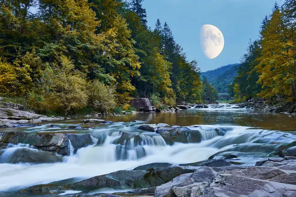 A fast mountain river flows over rocks in a forest, framed by autumn trees. The large moon rises above the mountainous horizon, casting a peaceful glow on the scene.