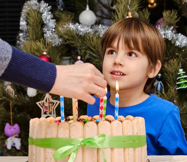 Genç bir çocuk, güzel dekore edilmiş bir doğum günü pastasında mum yakan, Noel ağacının arka planına kurulmuş, Noel neşesini Noel neşesiyle harmanlayan bir çocuğu hevesle izliyor..
