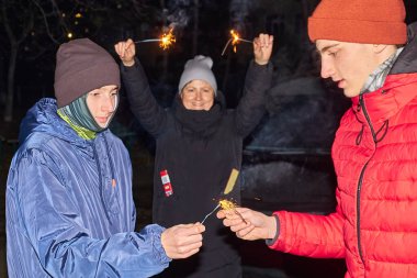 Geceleri havai fişeklerle kutlama yapan üç kişilik bir grup. İki genç adam maytap tutarken, arka planda bir kadın sevinçle kollarını kaldırıyor, o da bir maytap tutuyor..