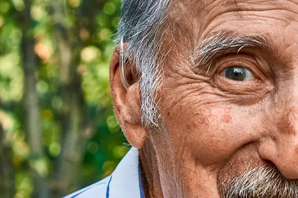 İşitme cihazı olan yaşlı bir adamın yakın plan portresi. Gözünü alıyor ve gülümsüyor. Fotoğraf, bulanık doğal bir arka planda geçen yaş, deneyim ve keyfi ön plana çıkarıyor..