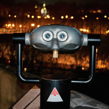 Halka açık bir dürbün izleyicisi, gece şehrin ışıklarıyla aydınlanan ön planda duruyor. İzleyicinin tasarımı bir yüzü andırıyor, şehir ortamında neşeli ve ilginç bir atmosfer yaratıyor..