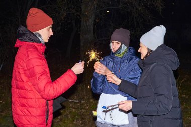 Geceleri havai fişeklerle kutlama yapan üç kişilik bir grup. İki genç adam maytap tutarken, arka planda bir kadın sevinçle kollarını kaldırıyor, o da bir maytap tutuyor..