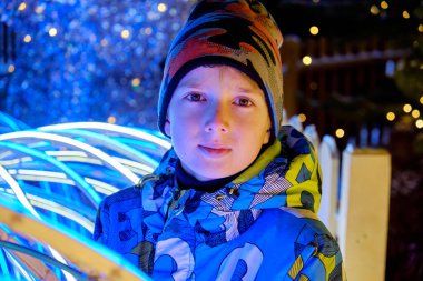 Kışlık ceket ve şapka giymiş bir çocuk gece parlak, renkli ışıklarla aydınlatılıyor. Arka planda Bokeh efektleri var. Sihirli bir tatil atmosferi yaratıyor..