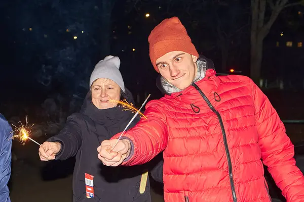 Geceleri havai fişeklerle kutlama yapan üç kişilik bir grup. İki genç adam maytap tutarken, arka planda bir kadın sevinçle kollarını kaldırıyor, o da bir maytap tutuyor..