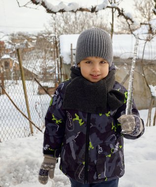 Sıcak kış kıyafetleri giymiş genç bir çocuk karlı bir ortamda açık havada gülümserken büyük bir buz saçağı tutuyor. Güneş ışığı buzdan yansıyor, soğuk ve güneşli bir günde neşeli anı yakalıyor..