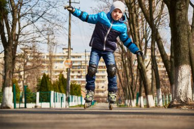 Parkta paten kayan, mavi ceketli, diz vatkalı ve eldivenli genç bir çocuk. Hareketin ortasında dengede, açık havada aktif bir günün tadını çıkarıyor..