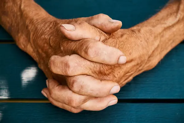 Yaş, bilgelik ve deneyim aktaran yapıyı, kırışıklıkları ve ayrıntıları vurgulayan sıkıca bağlanmış yaşlı ellerin yakın çekimi. Eller mavi bir arkaplanda duruyor, çarpıcı bir kontrast oluşturuyor..