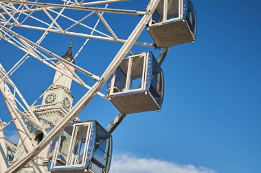 Dönme dolap kabinine yakın plan, arka planda açık mavi gökyüzüne karşı bir saat kulesi, mimari ve eğlence binasının bir karışımını yakalıyor..