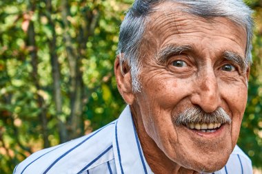 Elinde işitme cihazı olan mutlu yaşlı bir adamın portresi, dışarıda sıcak bir şekilde gülümsüyor. Parlak gözleri ve anlamlı yüzü doğayla çevrili. Yaşamın getirdiği neşe ve bilgeliği yansıtıyor..
