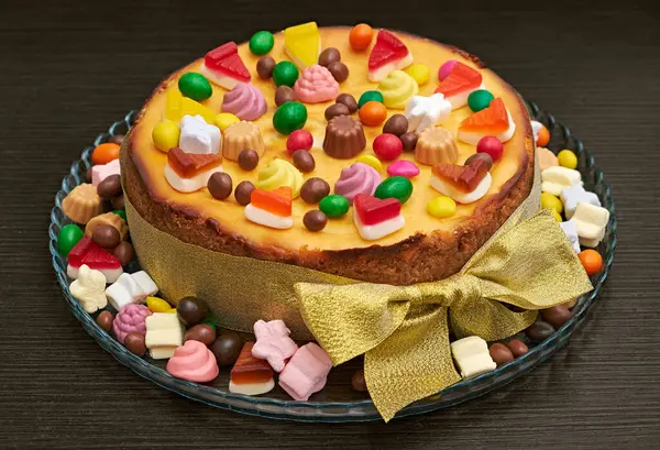 Renkli şekerler, jöle ve çikolatayla süslenmiş, altın bir fiyonklu cam bir tepsiye konmuş neşeli bir cheesecake. Kutlamalar ve tatlıya düşkünlük için mükemmel..