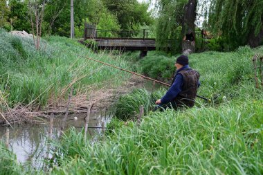 Sıcak giysili yaşlı bir adam küçük, fazla gelişmiş bir nehrin kenarında oturur ve geleneksel bir ağla balık tutar. Yeşiller yemyeşil çimenler ve sazlıklar etrafını sarmış, arkasında ahşap bir köprü var..