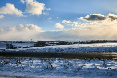 Yuvarlanan karla kaplı tarlalar, sahneyi kesen bir yol ve ufuk boyunca uzanan elektrik hatları, dağınık mavi bulutlu parlak bir gökyüzü altında..