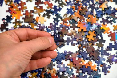 Bulmaca parçasını tutan bir elin yakın çekimi dağınık yapboz parçalarının bulanık arka planına karşı. Problem çözmenin, stratejinin ve yaratıcılığın sembolü. Takım çalışması, mantık ve beyin egzersizi için mükemmel..