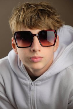 Aşırı büyük güneş gözlüğü takan, beyaz kapüşonlu, kendine güvenen genç bir çocuk stüdyoda poz veriyor. Modern estetiğe ve tarafsız bir geçmişe sahip moda portresi.
