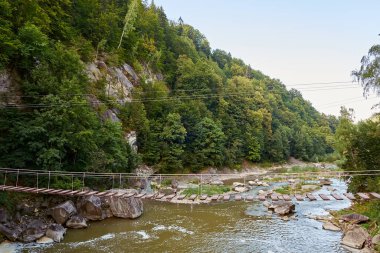 Ahşap tahtalardan yapılmış, yemyeşil orman ve kayalık uçurumlarla çevrili, hızlı akan bir dağ nehrinin üzerine uzanan, rustik asma köprüsü. Manzaralı ve maceralı bir doğa manzarası..