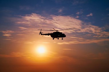 Gün batımında çarpıcı bir gökyüzüne doğru uçan askeri helikopterin silueti. Uçak havada süzülerek, arkasında ılık renkler ve yumuşak bulutlar olan dramatik bir sahne yaratıyor..