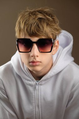 Aşırı büyük güneş gözlüğü takan, beyaz kapüşonlu, kendine güvenen genç bir çocuk stüdyoda poz veriyor. Modern estetiğe ve tarafsız bir geçmişe sahip moda portresi.