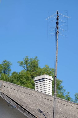 Eski televizyon anteni, tuğladan bacası olan bir kırsal evin üstündeki tahta bir direğe monte edilmiş. Mavi gökyüzü ve arka planda yeşil ağaçlar. Kırsal bir ortamda klasik teknoloji kavramı.