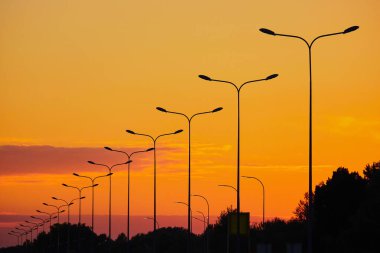 Canlı turuncu günbatımı gökyüzüne karşı modern sokak lambaları silueti. Şehir altyapısı bu dramatik akşam sahnesinde doğayla kesişiyor ve görsel olarak çarpıcı bir kontrast oluşturuyor..