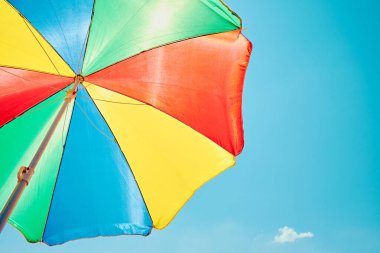 Güneşli bir yaz gününde açık mavi gökyüzüne karşı aşağıdan renkli bir plaj şemsiyesi. Rahatlamayı, tatili ve deniz kenarındaki eğlenceyi sembolize eden canlı ve neşeli bir sahne..