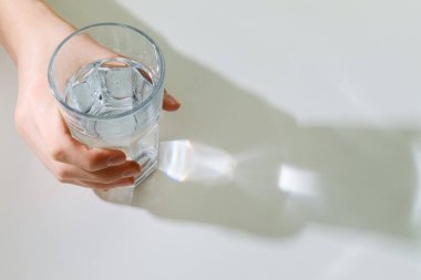 Beyaz bir masanın üzerinde buz küpleri olan bir bardak taze suyu tutan bir el. Güneş ışığı güzel yansımalar ve gölgeler yaratır. Minimalist, temiz ve ferahlatıcı bir kavram..