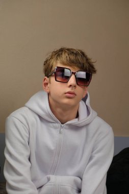 Aşırı büyük güneş gözlüğü takan, beyaz kapüşonlu, kendine güvenen genç bir çocuk stüdyoda poz veriyor. Modern estetiğe ve tarafsız bir geçmişe sahip moda portresi.
