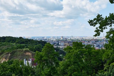 2025-15-06, Kyiv, Ukrayna. Yeşil tepeler ve bulutlu mavi gökyüzünün altındaki ağaçlarla çevrili modern şehir siluetinin manzaralı manzarası. Yaz mevsiminde kentsel ve doğa zıtlığı.