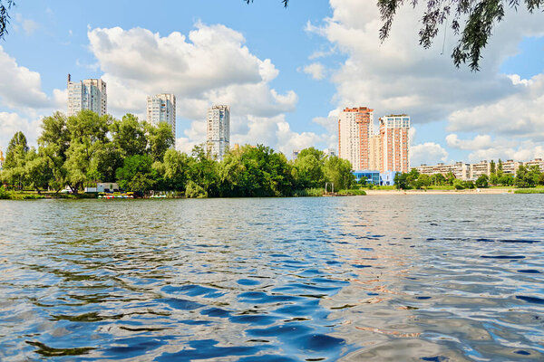 Красивый вид на озеро Тельбин в Киеве, Украина, с спокойной водой, пышной зеленью и современными жилыми многоэтажками под ярким голубым небом с облаками.