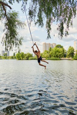 Genç bir adam bir yaz günü ağaca bağlı bir halatla suyun üzerinde sallanırken, arka planda şehir binalarıyla birlikte göle atlamaya hazırlanıyor. Şehir macerası ve açık hava eğlencesi.
