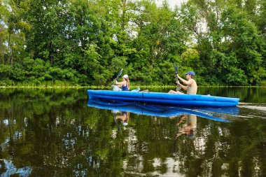 Yeşil ormanlarla çevrili sakin bir nehirde mavi bir teknede kano süren çift. Yaz macerası, aktif yaşam tarzı ve doğada su eğlencesi..