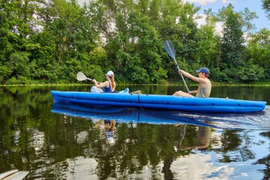 Yeşil ormanlarla çevrili sakin bir nehirde mavi bir teknede kano süren çift. Yaz macerası, aktif yaşam tarzı ve doğada su eğlencesi..