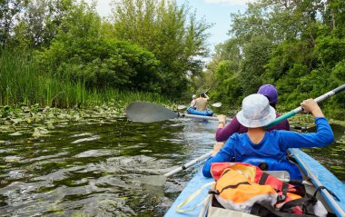 İnsanlar yemyeşil ağaçlarla çevrili sakin bir nehirde kano yapıyorlar. Yaz doğasında macera ve açık hava eğlencesi.