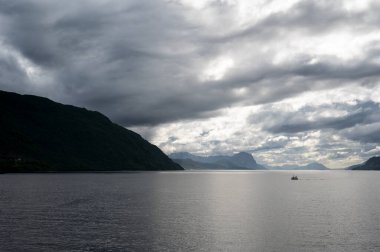 Norveç fiyortlarında yelken açan bir tekne. Bulutlu gökyüzü. Yalnızlık, kasvetli renkler.