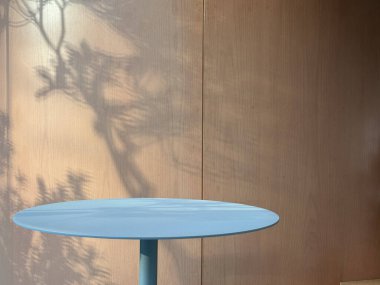 Ahşap bir duvarın önünde basit mavi yuvarlak bir masa, doğal gölgeler, sükunet ve iç estetik sergiliyor..