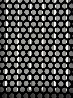 Eşit aralıklı dairesel deliklerle delik deşik edilmiş bir metal levhanın geometrik yakın çekimi arkasında dikey ışık demetlerini ortaya çıkarıyor. Siyah ve beyaz arasındaki güçlü zıtlık modern ve soyut bir mimari doku yaratır, arka plan veya de