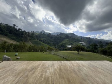 Geniş ahşap bir teras yemyeşil bir vadiye ve ağaçlar ve tarım arazileriyle kaplı yuvarlanan tepelere bakar. Yoğun bulutlar ve güneş ışınları ile dramatik gökyüzü barışçıl ve atmosferik bir doğa sahnesi yaratır. Model ya da tatil alanı için idealdir..
