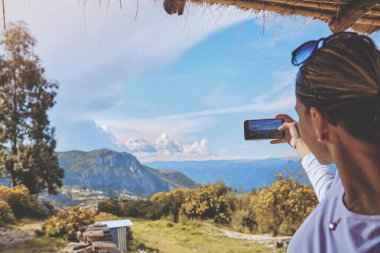 Kadın, dağ başında kulübede gün batımının keyfini çıkarırken kadın, denizin üstünde güneşin doğuşunu, tepeleri fotoğraflıyor.