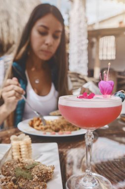 Bir kadın restoranda yemek yiyor ve üstünde çiçek olan pembe bir içecek var.