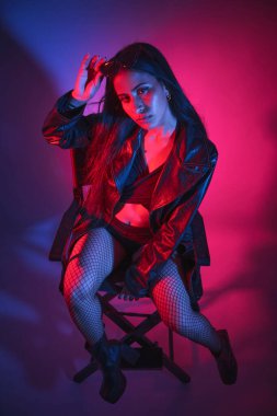 Siyah ceketli ve siyah çoraplı bir kadın sandalyede oturuyor. Siyah bir sütyen takıyor ve saçını arkaya çekiyor. Görüntüde karanlık ve kasvetli bir atmosfer var.