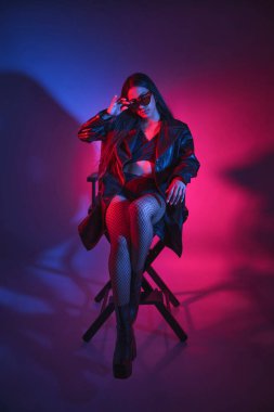 Siyah ceketli ve siyah çoraplı bir kadın sandalyede oturuyor. O, güneş gözlüğü ve siyah bir sutyen takıyor. Kadın kıyafetleri ile görüntüde biraz huysuzluk ve gerginlik var.