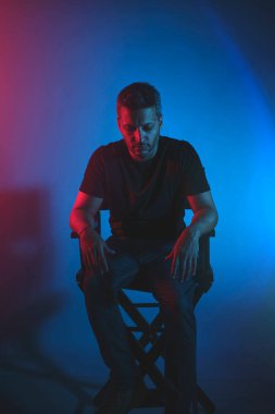 Bir adam elleri dizlerinin üzerinde bir sandalyede oturuyor. Sandalye siyah ve arka plan mavi. Adam derin düşüncelere dalmış