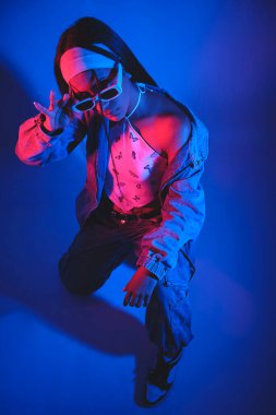 Kafasında güneş gözlüğü olan siyah beyaz elbiseli bir kadın fotoğraf için poz veriyor. Fotoğraf mavi ve mor renkte.