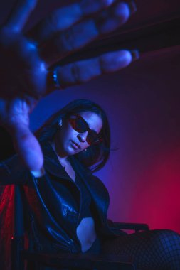 Siyah ceketli ve siyah pantolonlu bir kadın sandalyede oturuyor. O, güneş gözlüğü ve siyah bir sutyen takıyor. Görüntüde tuhaf ve gizemli bir his var.