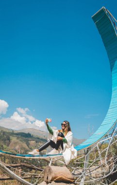 Mavi bir yapının üzerinde oturan bir kadın selfie çekiyor. Yapı mavi ve kavisli bir tasarımı var. Gökyüzü açık ve mavidir ve gökyüzünde hiç bulut yoktur.