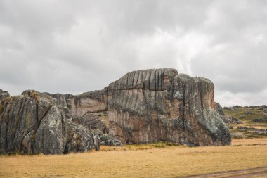 Ortasında küçük bir delik olan büyük bir kaya oluşumu. Gökyüzü bulutlu ve çimenler kuru.
