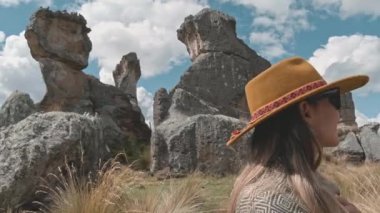 Şapka ve mavi yelek giyen bir kadın tarlada bir kayanın üzerinde oturuyor. Gökyüzünü işaret ediyor.