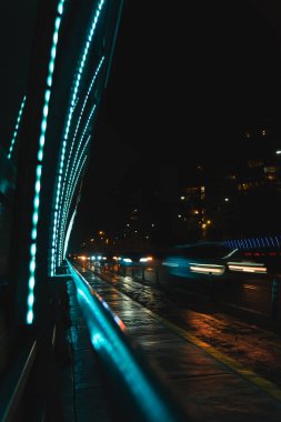Geceleri arka planda yeşil ışıklı bir köprü olan bir şehir caddesi. Sokaklar ıslak ve ışıklar açık.